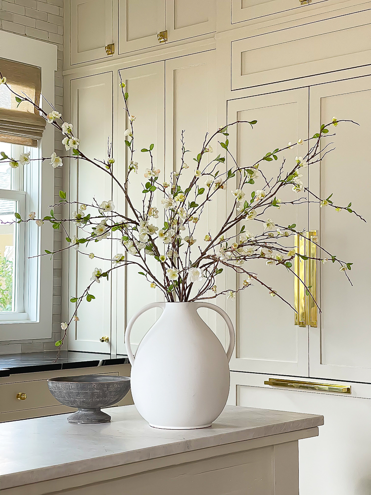 faux cream blossom branch or quince branch in white jug vase with handles and cream cabinets in background 